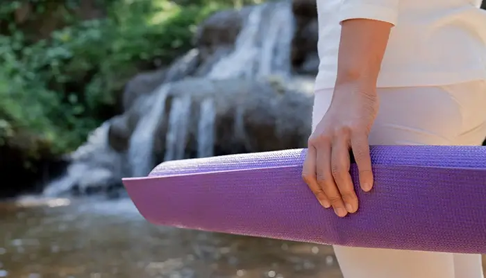 Introspective image featuring a woman holding a yoga mat in front of a beautiful, serene scene outdoors in front of a small natural waterfal